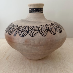 May include: A light tan, rounded ceramic vase with a narrow neck and a wide, bulbous body. The vase features hand-painted black geometric patterns, including a band of repeating oval shapes around the shoulder and a cross-hatch design on the neck.
