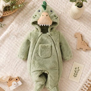 Peut inclure: Une combinaison une pièce en peluche vert sauge dotée d'une capuche en forme de tête de dinosaure avec un museau beige, des dents blanches et une petite crête verte. Le vêtement possède des manches longues, des pieds intégrés et une poche plaquée.