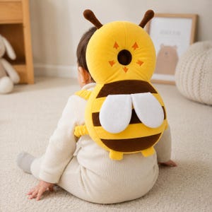 May include: A yellow and brown bee-shaped head and back protector cushion with white wings, two antennae, and elastic shoulder straps. The cushion features a central circular mesh cutout and orange arrow accents on the yellow head portion.