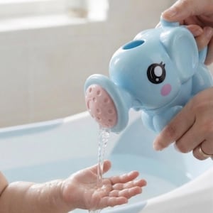 Peut inclure: Un arrosoir en plastique bleu clair en forme d'éléphant avec un embout rose perforé. Le jouet présente de grands yeux noirs et blancs ainsi que des joues roses circulaires. L'eau s'écoule du bec vers une petite main, montrant sa fonction.