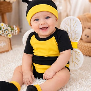 Peut inclure: Un body à manches courtes rayé noir et jaune avec des ailes blanches attachées ornées de broderies dorées, accompagné d'un bonnet noir et jaune assorti avec deux antennes et de chaussettes noires à revers jaunes portés par un nourrisson.