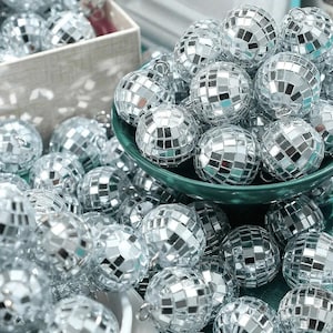 May include: A collection of small, spherical disco balls covered in tiny, reflective silver mirror tiles. Each ball features a small metal loop at the top for hanging. The disco balls are piled together in a dark green bowl and scattered around.
