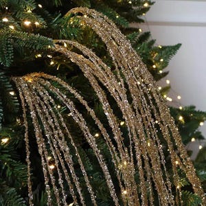 Peut inclure: Un ensemble de longues tiges décoratives flexibles composées de multiples fils métalliques dorés recouverts de petites perles réfléchissantes transparentes et dorées qui retombent en cascade, créant un aspect texturé scintillant.