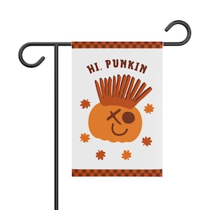 Könnte beinhalten: Eine weiße Gartenflagge mit einer zentralen orangefarbenen Kürbisillustration, die einen braunen Irokesenschnitt, ein X-förmiges Auge und ein kreisförmiges Auge aufweist. Der Text HI. PUNKIN steht darüber, umgeben von kleinen orangefarbenen Ahornblättern und braunen Karorändern.