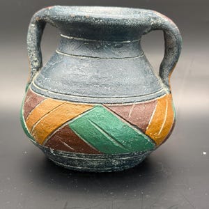 May include: A small, round, two-handled ceramic vessel with a textured, dark gray finish. The lower portion features a geometric pattern of diagonal stripes in shades of green, brown, and orange, separated by incised lines on the surface.