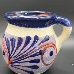 May include: A round ceramic mug with a cream-colored base, featuring a wide dark blue rim and a matching handle. The body is decorated with a light purple band painted with dark blue petal-like patterns and thin orange circular accents.