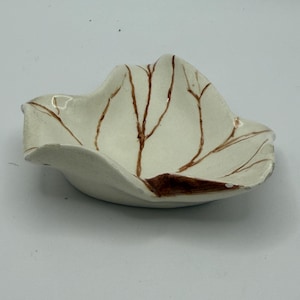 May include: A small, off-white ceramic dish with an irregular, wavy rim. The interior surface features a hand-painted brown design resembling thin, branching tree veins or leaf patterns, with a solid brown accent near the base of the rim.
