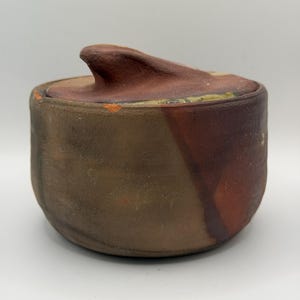 May include: A cylindrical ceramic jar with a lid featuring a prominent, curved handle. The exterior displays a textured, matte finish in earthy tones of brown, charcoal, and deep red, with a visible chip on the upper rim of the base.
