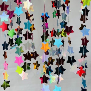 May include: A hanging decoration consisting of multiple vertical strands of reflective, holographic stars in various colors including red, blue, green, yellow, and silver. Each star is connected by a thin, light-colored string, creating a cascading effect.