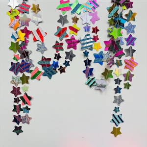 May include: A decorative garland featuring numerous star-shaped cutouts in various sizes and colors, including metallic, glittery, and striped patterns. The stars are connected by thin white thread, creating a vertical hanging display.