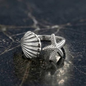 Può includere: Un anello aperto color argento con una conchiglia testurizzata su un'estremità e una stella marina a cinque punte sull'altra. La fascia in metallo è lucida e riflettente, mostrando creste dettagliate sulla conchiglia e piccoli punti in rilievo.