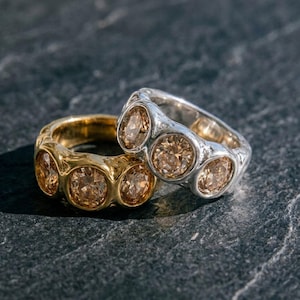 Op de afbeelding: Twee ringen, één goudkleurig en één zilverkleurig, elk voorzien van drie ronde, lichtbruine gefacetteerde stenen gezet in een dikke, getextureerde metalen band. De ringen liggen over elkaar heen en tonen de afwerking en de stenen.