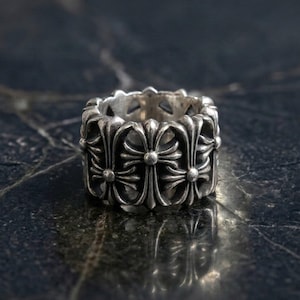 May include: A wide silver ring featuring a repeating pattern of embossed cross motifs around the entire band. The metal has a polished finish with darkened recessed areas that highlight the intricate details of the cross designs.