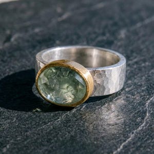 Op de afbeelding: Een zilveren ring met een brede, gehamerde band en een centrale ovaalvormige, gefacetteerde lichtgroene edelsteen gezet in een gladde, gepolijste goudkleurige zetting. De ring toont een contrast tussen het gehamerde zilver en het goud.