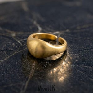 May include: A gold-toned ring with a smooth, rounded, and slightly domed surface. The band features an open design, allowing for adjustment. The metal has a brushed, matte finish that catches the light, creating a soft glow across the curved top.