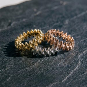 May include: Three metal rings featuring a continuous band of pointed, pyramid-shaped studs. The set includes one gold-toned ring, one rose gold-toned ring, and one silver-toned ring, all arranged in a cluster on a dark, textured surface.