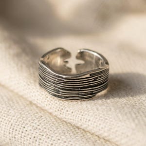 May include: A silver-toned open ring featuring a textured surface with horizontal, irregular grooves and lines. The band has a distinct gap at the top, showcasing a metallic finish with darkened recessed areas that emphasize the linear pattern.
