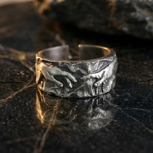 May include: A silver-toned metal ring featuring a textured, rugged surface with a raised relief design depicting two hands reaching toward each other. The band has an open-ended construction, showcasing a weathered, oxidized finish throughout.