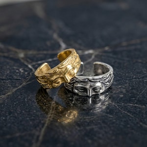 May include: Two open-band rings featuring a sculpted human face design with flowing hair details. One ring is finished in a polished gold tone, while the other is finished in a silver tone with darkened recessed areas to highlight the facial features.