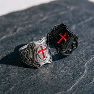 Könnte beinhalten: Zwei schildförmige Ringe, einer aus poliertem silberfarbenem Metall und einer aus mattem schwarzem Metall. Beide Ringe zeigen ein zentrales, erhabenes Schildelement mit einem leuchtend roten Kreuz und dekorativen Verzierungen an den Seiten.