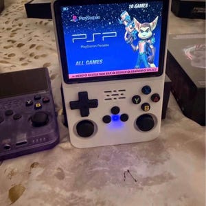 May include: A white handheld gaming console with a vertical screen displaying a PlayStation Portable menu interface featuring Ratchet. The device includes a black directional pad, two black analogue sticks, four coloured action buttons labelled A, B, X, Y, and three small central function buttons with a blue light.