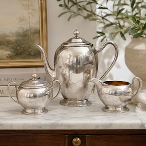 May include: A silver tea service set consisting of a tall teapot with a lid, a lidded sugar bowl with two handles, and a creamer with a gold-toned interior. Each piece features a polished metallic finish and a decorative base with intricate detailing.
