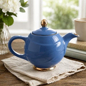 May include: A glossy blue ceramic teapot featuring a rounded body, a curved handle, and a short spout. The lid is topped with a small, polished gold-tone knob, and the base of the teapot is finished with a matching gold-tone metallic trim.