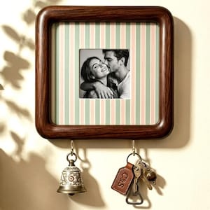 Pode incluir: Uma moldura quadrada de madeira escura com cantos arredondados e um passe-partout com riscas verticais verde-claras e cor-de-rosa. A moldura contém uma fotografia a preto e branco de duas pessoas. Dois ganchos de latão seguram um sino e um porta-chaves.