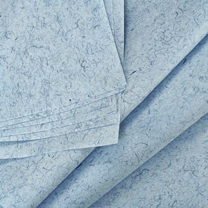 Könnte beinhalten: Ein Stapel hellblauer, handgeschöpfter Papierbögen mit einer strukturierten Oberfläche, in die dunkelblaue Fasereinschlüsse eingearbeitet sind, was den überlappenden rechteckigen Seiten ein natürliches und organisches Aussehen verleiht.