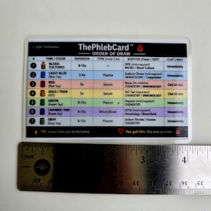 May include: A rectangular reference card titled ThePhlebCard Order of Draw, featuring a table with seven rows of color-coded blood collection tube information. A metal ruler is positioned below the card, showing a width of approximately 4.5 inches.
