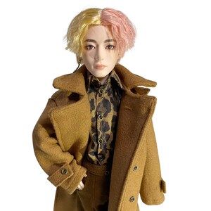 May include: A doll with split-dyed yellow and pink hair, wearing a brown trench coat over a brown and black leopard print button-down shirt. The doll has a neutral facial expression, defined eyebrows, and a small silver chain earring on the right side.