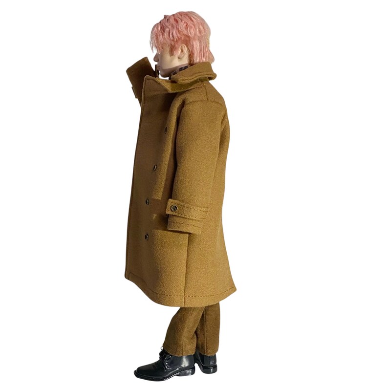 May include: A collectible doll with short pink hair wearing a tan double-breasted wool-style coat with a wide collar, matching tan trousers, and black lace-up boots. The coat features visible buttons down the front and on the sleeve cuffs.