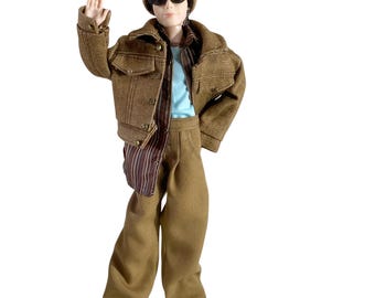 May include: A doll wearing a tan beanie with a "Hand made" label, black cat-eye sunglasses, a brown denim jacket, a brown and white striped button-down shirt, a light blue undershirt, and tan trousers. The doll has one arm raised in a waving gesture.