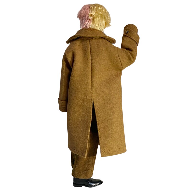 May include: A doll with short, two-toned pink and blonde hair, wearing a long, tan-colored wool-like coat with a wide collar, long sleeves with buttoned cuffs, and a center back vent, paired with matching tan trousers and black dress shoes.