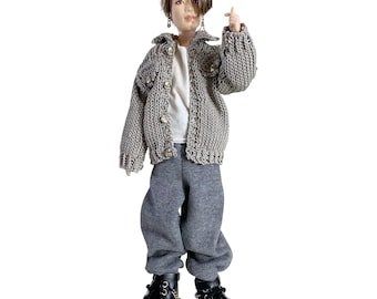 May include: A doll with brown hair wearing dark sunglasses, a grey knit jacket with buttons, a white shirt, grey trousers, and black boots. The doll has one hand raised in a finger heart gesture and wears small dangling earrings on its ears.