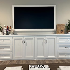 May include: A white media console with six drawers and two central cabinet doors featuring silver hardware. A large television with a white frame is mounted above. The console is decorated with small evergreen trees and nutcracker figurines.