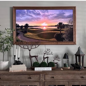 May include: A framed landscape print of a sunset over a golf course with palm trees and a winding path. Below, three metal deer silhouettes, a small sign reading I LOVE US, a black pig figurine, a lantern, and a lamp sit on a wooden console table.