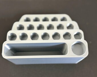 May include: A light gray 3D printed organizer with a tiered design. The top three levels feature rows of circular holes for storage, while the bottom level contains a wide rectangular slot and a single circular compartment on the right side.