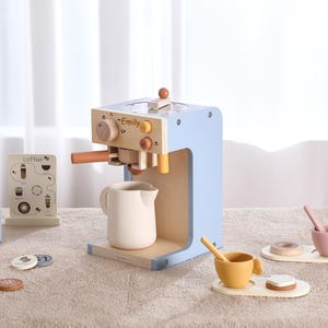 Könnte beinhalten: Eine Spielzeug-Espressomaschine aus Holz in Hellblau und Naturholztönen mit dem Namen Emily auf der Vorderseite. Sie enthält ein weißes Kännchen, eine kleine Menükarte, einen blauen Milchkarton sowie zwei Tassen mit Löffeln und kleinen Leckereien.