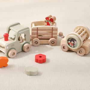 May include: A wooden toy train set featuring a sage green engine with the name Noah printed on the side, a cargo wagon with a squirrel figure, and a carriage with a rotating drum. Two loose wooden shapes, one orange and one red, sit nearby.