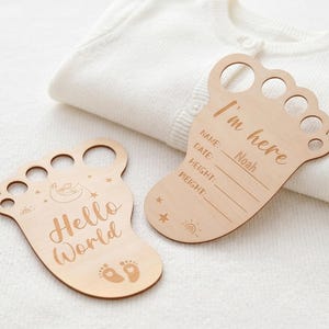 Op de afbeelding: Twee lichtgekleurde houten voetvormige bordjes voor pasgeborenen. Eén bordje bevat de tekst Hello World met een maan en sterren. Het andere bordje bevat de tekst I'm here met lijnen voor Name, Date, Height en Weight, met de naam Noah erin gegraveerd.
