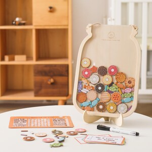 Op de afbeelding: Een lichtgekleurd houten bord in de vorm van een pot met een doorzichtig voorpaneel met diverse kleine houten fiches in de vorm van donuts en snoepjes. Een oranje rechthoekige checklist, een zwarte stift en een stapeltje kaarten liggen ervoor.