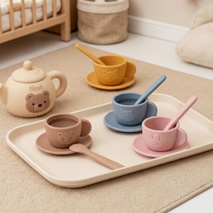Peut inclure: Un service à thé en silicone comprenant une théière beige avec un visage d'ours et le nom Noah, trois tasses avec soucoupes et cuillères en jaune moutarde, bleu et rose, et une tasse brune avec soucoupe et cuillère sur un plateau beige.