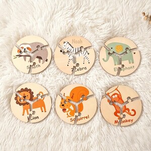 Op de afbeelding: Zes ronde houten puzzelstukken met illustraties van een luiaard, zebra, olifant, leeuw, eekhoorn en aap. Elk stuk is verdeeld in drie segmenten en bevat de diernaam in zwarte letters, met de naam Noah op het zebrapuzzelstuk.