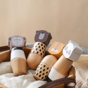 May include: A set of four wooden animal-themed stacking toys featuring a monkey, a bear, a tiger, and a panda. Each toy has a smooth, natural wood base, a painted midsection with geometric patterns or solid colors, and a stylized animal head topper.