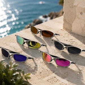 May include: Five pairs of sunglasses with metallic silver frames and distinct colored lenses, including brown, green, yellow, black, blue, and pink, arranged on a textured stone surface with a view of the ocean in the background.
