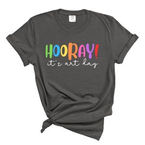 Puede incluir: Una camiseta gris oscuro con el texto "HOORAY! it's art day" en colores arcoíris.