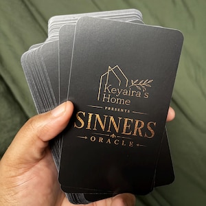 May include: A deck of rectangular oracle cards with a matte black finish. The top card features gold foil text that reads Keyaira's Home Presents SINNERS ORACLE, accompanied by a gold line drawing of a house outline and a small leafy branch.