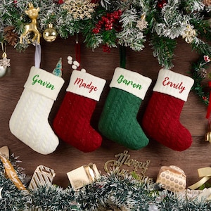 May include: Four cable-knit Christmas stockings hanging in a row. The first is white with the name Gavin in green, the second is red with Madge in red, the third is green with Baron in green, and the fourth is red with Emily in red text.