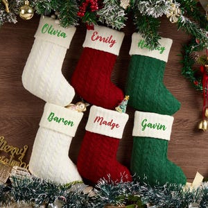 May include: Six cable-knit Christmas stockings in white, red, and green with white cuffs. Each stocking features embroidered names: Olivia, Emily, Harvey, Baron, Madge, and Gavin. The stockings are arranged in two rows of three against a wooden background.